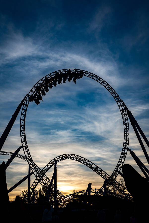 Sillhouette einer Achterbahn gegen den abendlichen blauen, mit zarten Wolken durchzogenen Himmel. Ein Zug der Achterbahn ist gerade in einem Looping.