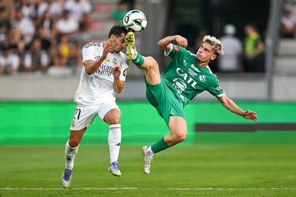 Szene aus einem Fussballspiel, ein Spieler in grünem Dress hat einen Fuß hoch in der Luft und berührt damit gleichzeitig Ball und Kopf eines Spielers in weißem Dress.