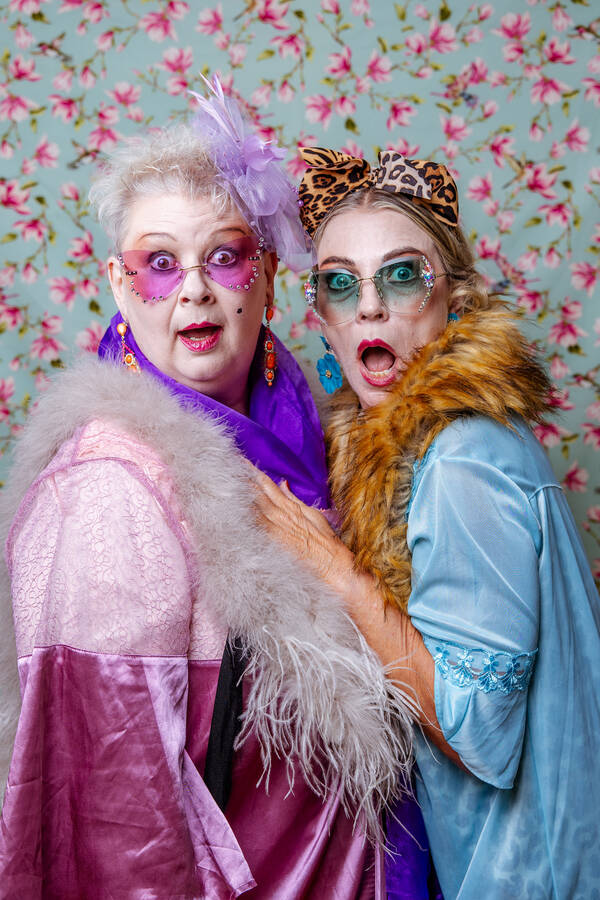 Zwei ältere Damen in knallig bunten Outfits mit bunten Brillen vor einer blumigen Tapete.