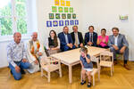 Um einen neuen Kindertisch sitzen einige Erwachsene, darunter Landeshauptmann Christoph Drexler und ein Pfarrer. Auf einem kleine Stuhl sitht ein kleines Mädchen und heult.