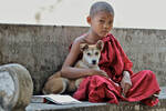 Ein Bub sitzt in Priesterkleidung in Myanmar und umarmt einen Hund. Neben ihm liegt ein dickes aufgeschlagenes Buch. 