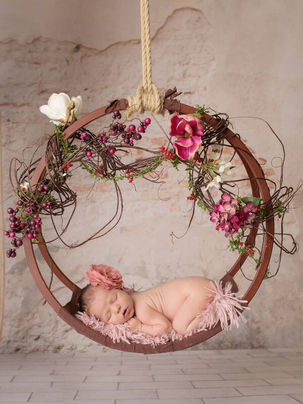 In einem hängenden, blütengeschmückten Reif liegt ein schlafendes Baby mit einer Blüte am Kopf.