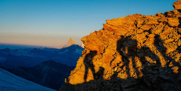 Sonnenaufgang in den schweizer Bergen. Im Hintergrund wird das Matterhorn von der Morgensonne erleuchtet. im Vordergrund zeichnet die Morgensonne die Schatten mehrerer Menschen auf einen Felsen.
