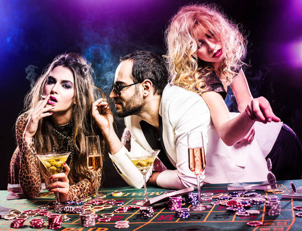 Ein Mann und zwei Frauen lehnen rauchend und Cocktail-trinkend an einem Roullett-Tisch.