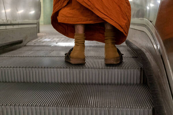 Detailaufnahme der Füße einer alten Frau in ockerfarbigen Socken und Schlapfen auf der obersten Stufe einer Rolltreppe.