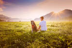 Jemand sitzt mit dem Rücken zum Betrachter neben einem Hund in der morgendlich Taunassen Wiese und genießt den Sonnenaufgang