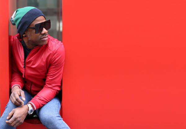 Ein dunkelhäutiger Mann mit Mütze und Sonnenbrille sitzt in einer engen Nosche eines roten Objektes.