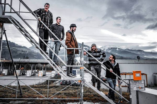 Fünf Männer stehen auf einer provisorischen Stiege eines Gerüstes. Im Hintergrundist eine Mautstelle und verschneite Berge. Auf einem Schild steht, dass das betreten des Gerüstes verboten ist.