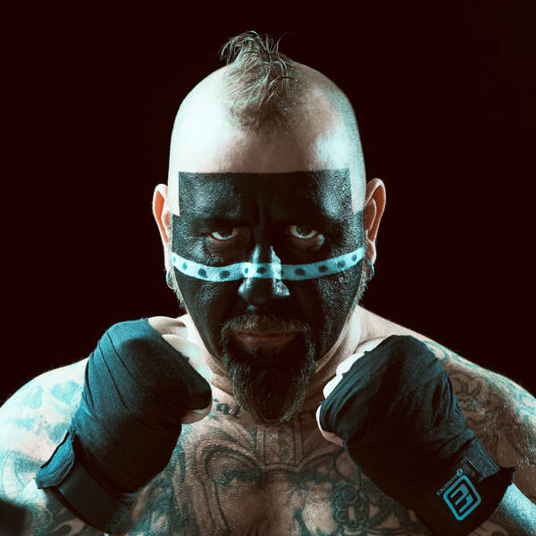 Ein Wrestler mit bandagierten Händen und Irokesen-Frisur. Das Gesicht ist schwarz geschminkt mit einem türkisen Band über die Nase.