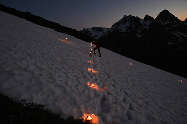Brennende Lichter im Schnee in den Bergen in der Abenddämmerung wie bei den Herzjesu-Feuern. Ein Mensch hebt sich am Kreuzungspunkt der Lichter dunkel vom Schneefeld ab.