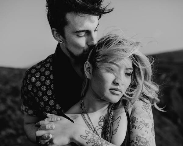Ein junger, dunkelhaariger Mann mit Bart steht hinter einer jungen Frau mit vielen Tatoos und Ketten. sie halten sich an einer Hand und er küsst ihren Kopf. Schwarz-Weiß.