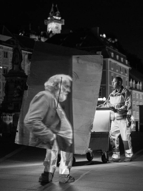 Nächtliche Aufnahme vom Hauptplatz in Graz. Mitarbeiter der Müllabfuhr bei der Arbeit, einer trägt einen großen Karton auf dem ein alter Mann zu sehen ist. Schwarz-Weiß