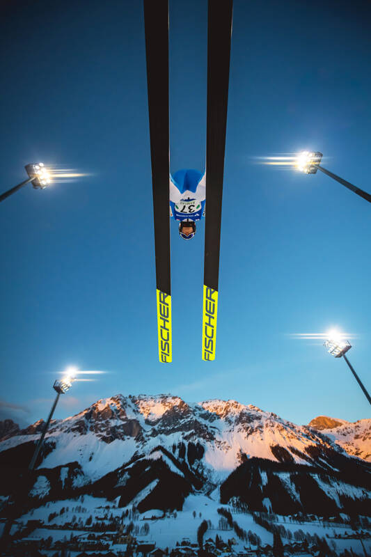 Schispringer in der Abenddämmerung mit Flutlicht direkt beim Absprung von unten fotografiert. Berge im Hintergrund.