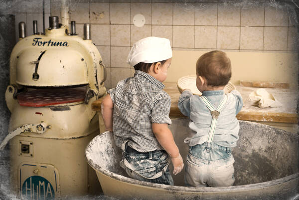Zwei Kinder stehen mit Mehl bestäubt in einer riesigen Teigschüssel in einer Bäckerei. Eines der Kinder trägt eine Bäckermütze.