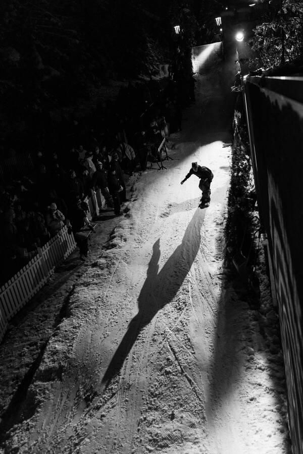 Ein Snowboarder fährt in der Dunkelheit, ein Scheinwerfer erzeugt einen ausgeprägten Schatten, in der Dunkelheit sind Zuschauer zu erkennen. Schwarz-Weiß.