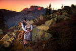 Ein junges Paar in Tracht und Lederhose stehen an einem grobblockigen Berghang gegen einen großen flechtenbewachsenen Stein gelehnt in sich versunken. Im Hintergrund beleuchtet die Abendsonne rotglühende Bergspitzen.