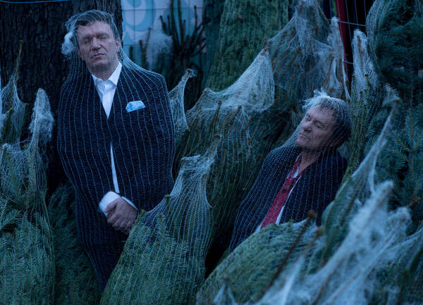 Der Pianist Markus Schirmer und der Schauspieler Wolfram Berger sind inmitten von verpackten Weihnachtsbäumen in die typischen Kunststoffnetzen für Christbäume verpackt.