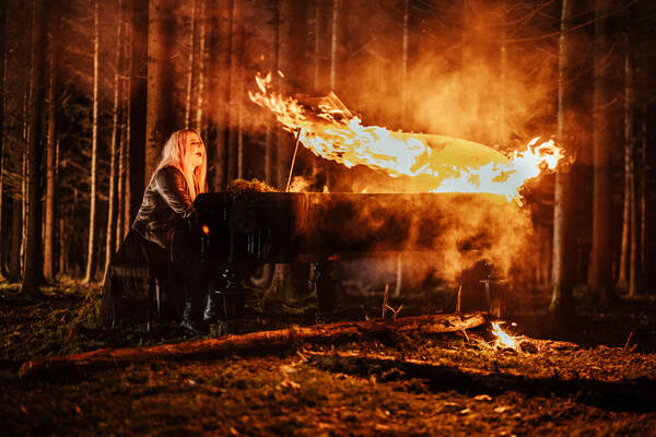 Eine Frau sitzt mitten im nächtlichen Wald an einem brennenden Klavier und spielt.