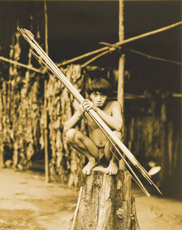Ein Mann des Yanomami-Volkes aus dem Amazonas mit Bogen und Pfeilen auf einem Baumstock. Im Hintergrund hölzerne Gerüste zum Trockenen von Lebensmitteln.