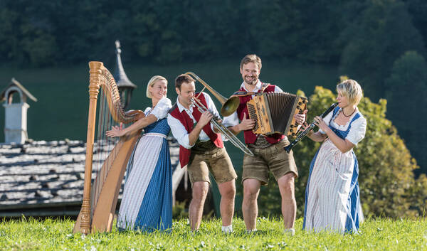 Vierköpfige Musikgruppe aus zwei Männern und zwei Frauen mit Harfe, Klarinette, Posaune und Zieharmonika vor einem Schindelgedeckten Bauernhaus mit Glockenturm.