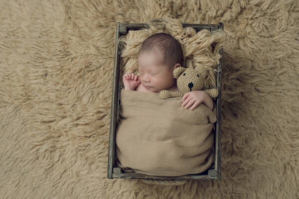 Ein Neugeborenes liegt in eine Leinendecke gewickelt mit einem gestrickten Bären in einer Holzkiste auf einem Schaffell.