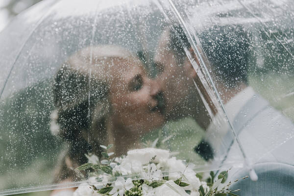 Portrait eines küssenden Brautpaares durch einen transparenten, regennassen Regenschirm.