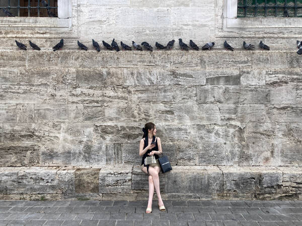 Eine Frau in sommerlicher Kleidung mit mehreren Shopping-Taschen sitzt auf einem Mauersims. auf einem Vorsprung über ihr sitzen zahlreiche Tauben.