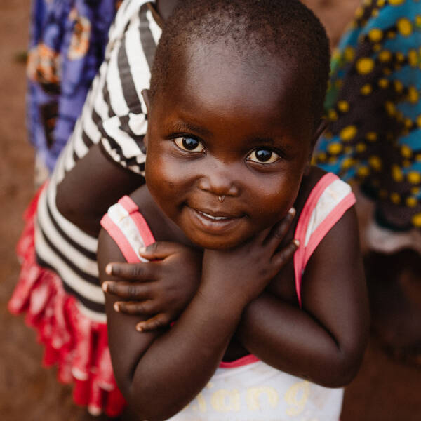 Ein dunkelhäutiges, kleines Mädchen blickt mit großen, dunklen Augen in denen sich der Himmel spiegelt nach oben in die Kamera. Sie hat die Arme vor der Brust verschränkt. Im Hintergrund sieht man andere Kinder in traditionellen afrikanischen Kleidern.