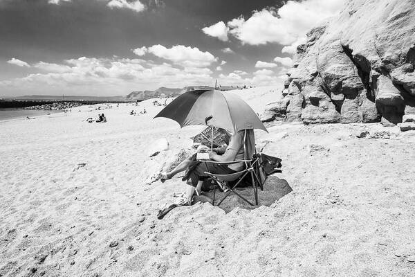 Ein Paar sitzt unter einem Schirm am sonnigen Strand, einer liest die Zeitung 
