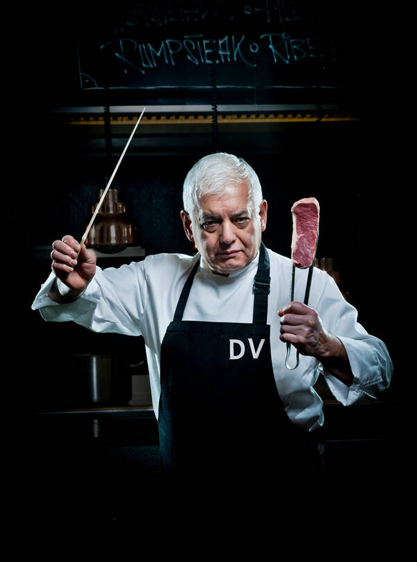 Portrait eines Kochs mit einem Dirigentenstab in der einen Hand und einer Zange mit einem Steak in der anderen Hand. Der Koch trägt eine weiße Weste und eine schwarze Schürze mit der aufschrift DV.