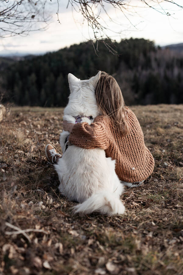 Ein großer weißer Hund sitzt neben einer Frau mit langem, blondem Haar, die sich an den Hund anlehnt und ihren Arm um ihn gelegt hat. Beide schauen in die Ferne.