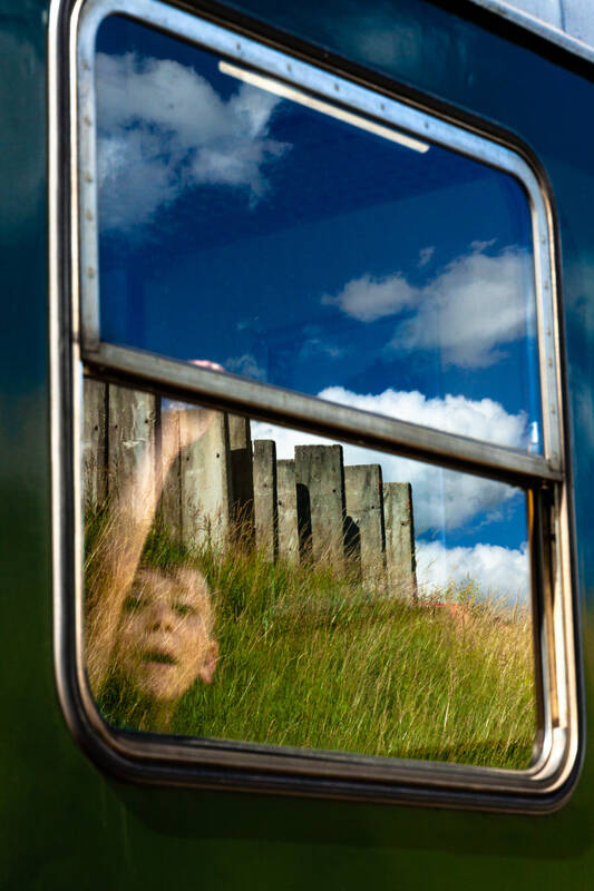 Blick von Außen durch ein Zugfenster auf das Gesicht eines Kindes. Im Fenster spiegelt sich die Landschaft.