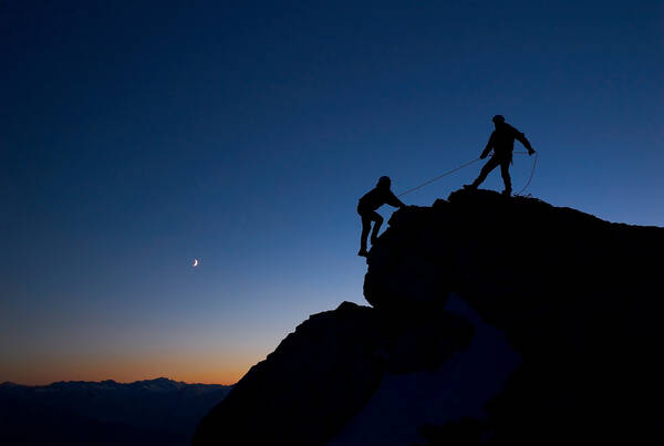 Silhouette zweier Bergsteiger gegen den abendlichen Himmel. Einer sichert von oben den nachkommenden an einem Seil.