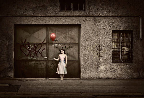 Ein Mädchen mit einem roten Ballon steht vor einem Garagentor eines graubraun heruntergekommenen Hauses. Graffiti auf Tor und Wänden.