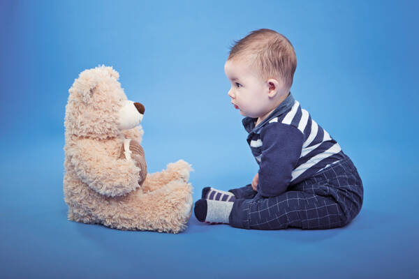 Ein Baby und ein Kuschelbär sitzen sich in einem blauen Raum gegenüber.