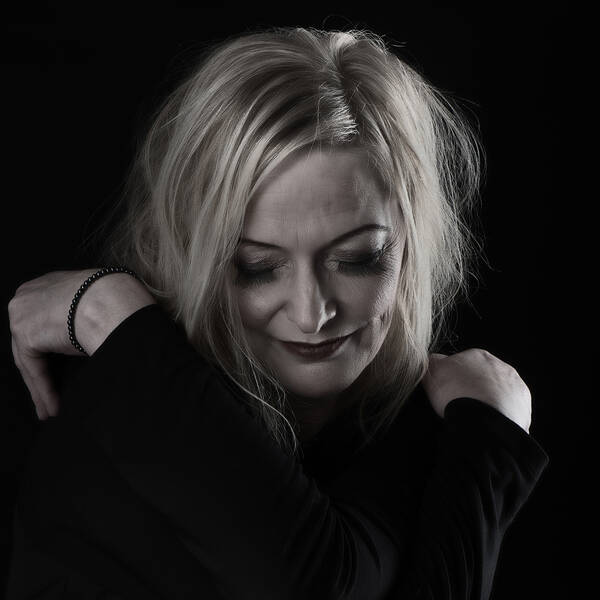 Eine blonde Frau in dunklem Gewand hat die Hände überkreuzt auf die eigenen Schultern gelegt. Low Key.