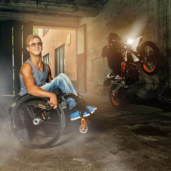 Ein Mann im Rollstuhl macht einen Wheelie. Im Hintergrund macht ein Motorradfahrer auch einen Wheelie.