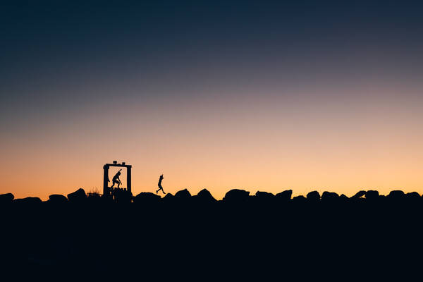 Vor einem abendlich verfärbten Himmel sieht man die Silhouette von Kindern, die über große Steinblöcke klettern.