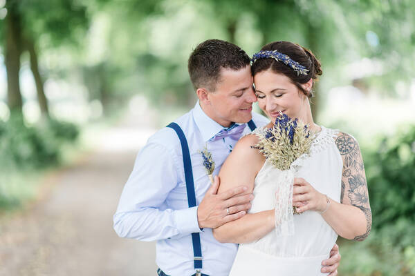 Ein Brautpaar auf einem Weg im Wald, er steht hinter ihr, sie legen die Köpfe aneinander, er hält sie an Arm und Hüfte.