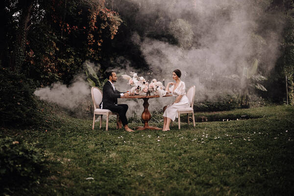 Ein elegant gekleidetes Paar sitzt an einem üppig gedeckten Tisch inmitten eines Gartens. Von hinten ziehen Nebelschleider durchs Bild.