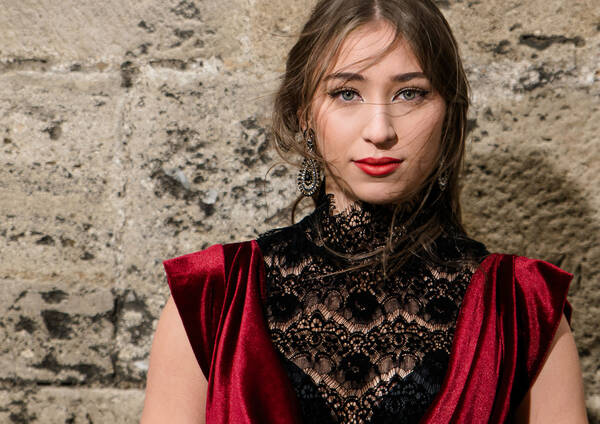 Portrait einer jungen Frau in elegantem, spitzenbesetztem Kleid in rot und schwarz vor einer Steinmauer.