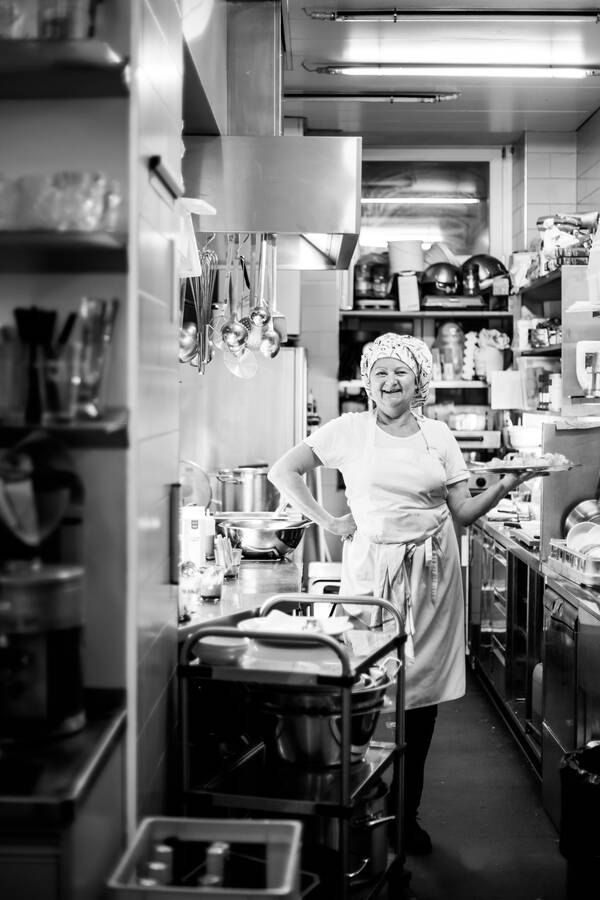 Eine ältere Frau steht mit einem Servier-Tablett in einer Restaurant-Küche. Schwarz-Weiß..