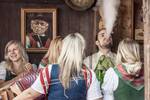 In einem Holzgetäfelten Gastzimmer sitzt der entführte Bräutigam mit jungen blonden Frauen an einem Tisch und bläst Rauch aus dem Mund. Eine der Frauen spielt Harmonika.