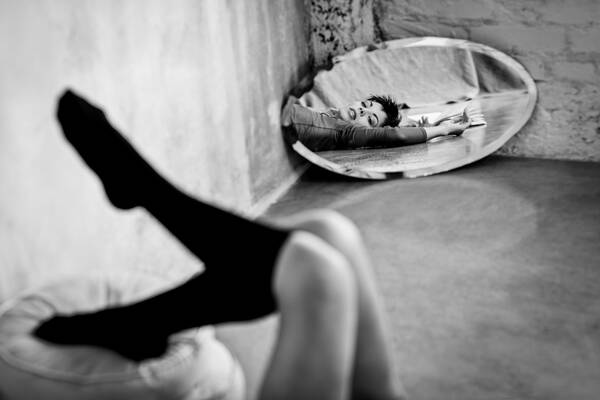 Ein ovaler Spiegel mit Facettenschliff steht in einer Ecke vor einer weiß getünchten Ziegelwand. Im Spiegel sieht man Brust, Kopf und Arme eines Models das am Boden liegt und mit gesenkten Lidern über den Spiegel in die Kamera sieht. Ein Buch liegt neben ihr. Im Vordergrund sieht man in der Unschärfe die Beine des Models mit dunklen Kniestrümpfen, die sich auf runden Pölstern abstützen. Schwarz-Weiß.