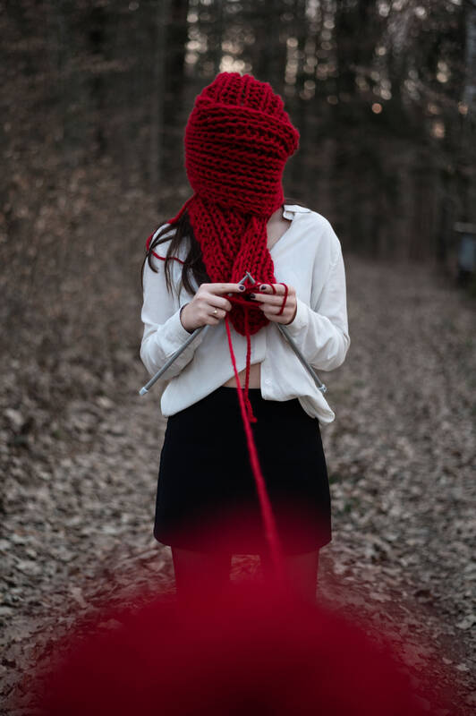 Eine junge Frau steht strickend auf einem Waldweg. Ihr Kopf ist in einen roten Strickschal eingewickelt, an dem sie gerade strickt und der Wollfaden läuft zur Kamera und am unteren Bildrand ist der rote Wollknäuel zu sehen.