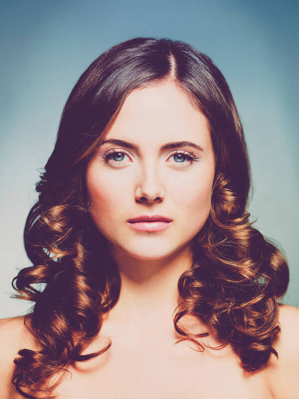 Portrait einer jungen Frau mit langen, brünetten, gelockten Haaren, die sie offen Trägt.