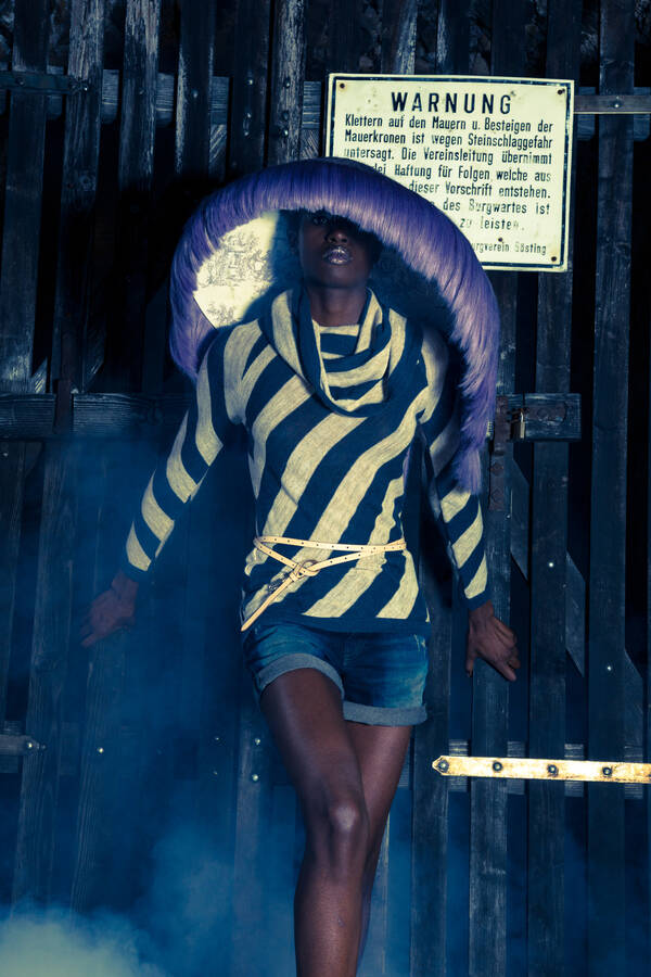 Eine Frau in Hotpants und einer schräg gestreiften blau-gelben Bluse mit einem überdimensional großen, ferderbesetzten Hut vor einem Holztor auf dem ein Warnschild angebracht ist.