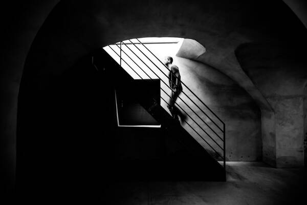 Ein Mann geht eine Treppe aus einem sehr dunklen, modernen, gewölbeartigen Raum nach oben, wo es heller wird. Schwarz-Weiß.