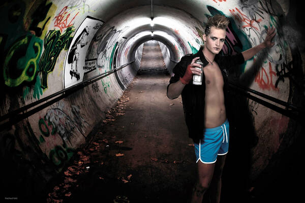Ein Junger Mann mit Punkfrisur in Shorts und offenem Hemd lehnt sich an eine Wand in einer Unterführung. Die Wände sind mit Graffitti besprüht. Der junge Mann hat eine Spraydose in der Hand.