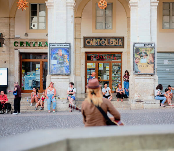 Straßenszene: Menschen sitzen auf den Stiegen vor Geschäften in einem Laubengang. Im Vordergrund sieht man unschaft eine Person mit Gitarre.